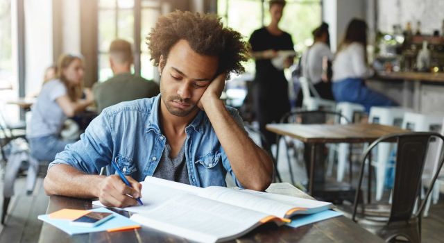 sad unhappy dark skinned bearded student feeling frustrated while preparing lessons college writing down his copybook with pen leaning elbow looking notes with upset expression